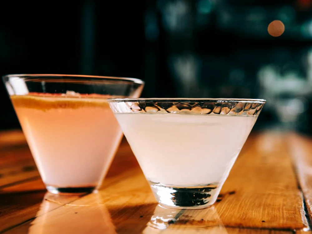 Two chilled coupe glasses on a warm wooden bar in low light